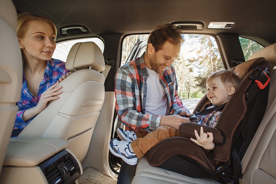 Personal Insurance - A Father is Buckling His Toddler Son into His Carseat in the Back of the Family Car While the Mother Watches on
