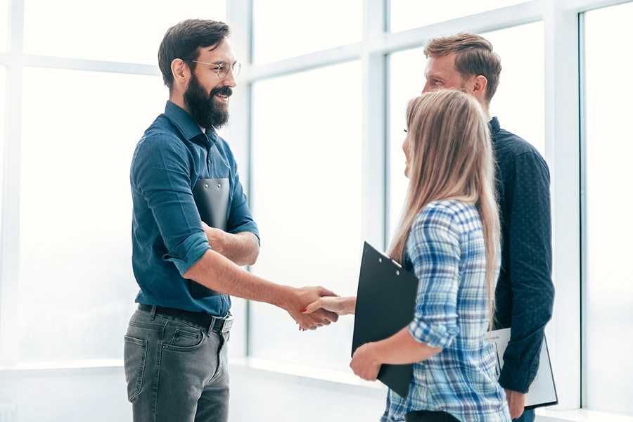 Refer a Friend - A Man With a Beard and Glasses is Shaking Hands With a Woman Holding a Clipboard While Another Man is Standing There With a Clipboard in an Office