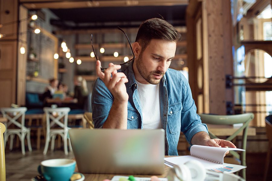 Resources - A Man is Sitting at a Table in a Cafe While Taking Notes and Using a Laptop