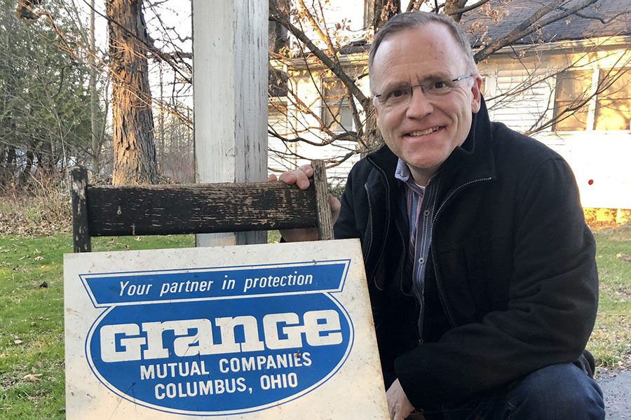 Homepage - Dan Reid Squatting Down by a Grange Insurance Sign With Trees and a House in the Background